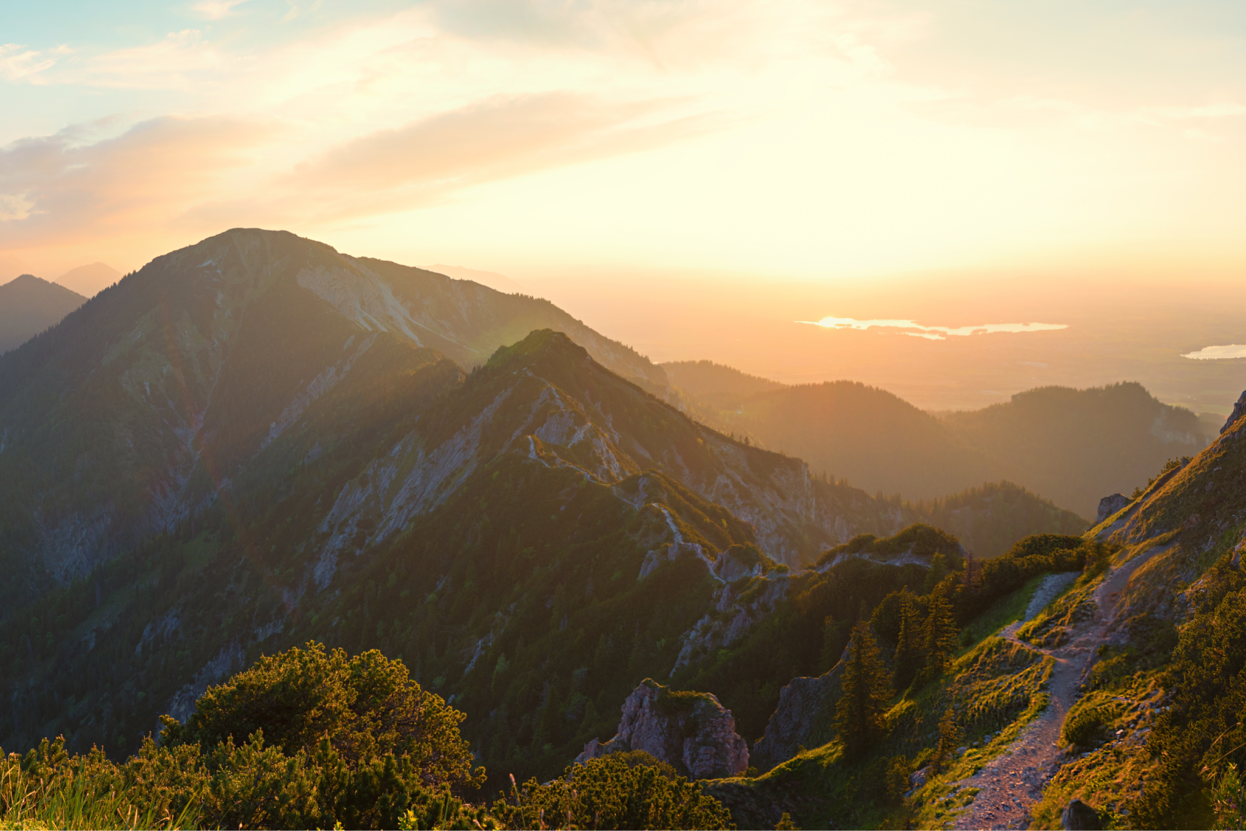 Sunset over a mountain ridge with rocky peaks, green trees, and a winding hiking trail.