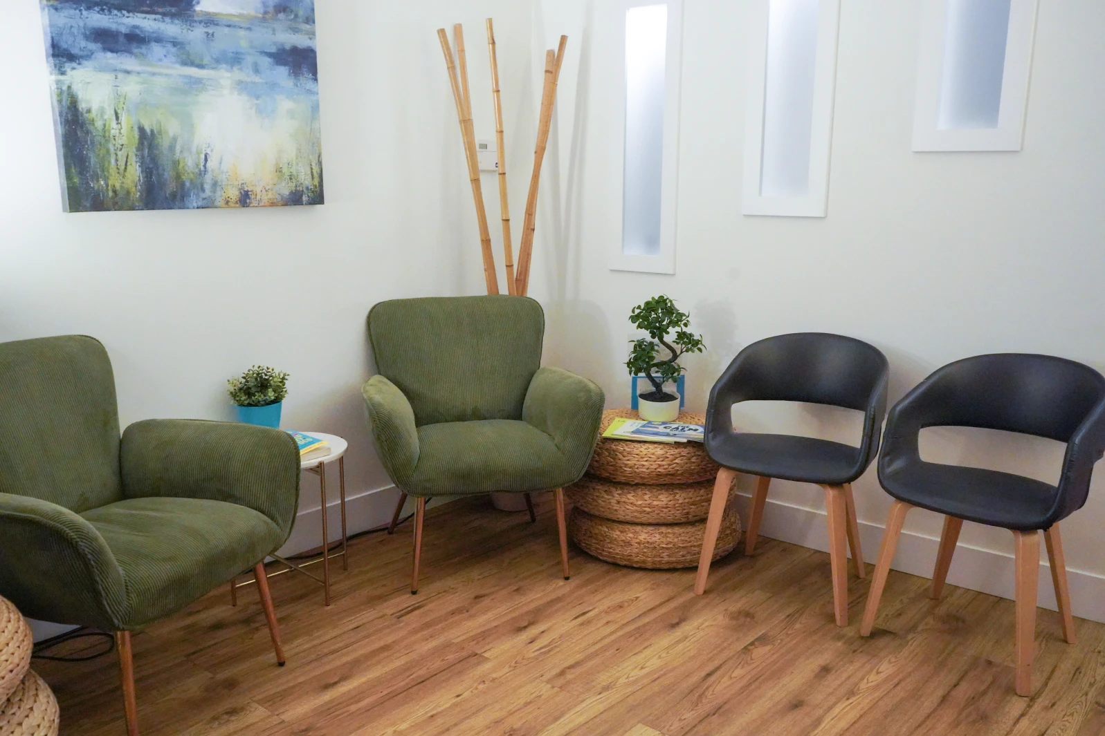 Cozy waiting room with two green armchairs, two black chairs, woven round tables, a small plant, bamboo sticks, and a landscape painting on a white wall.
