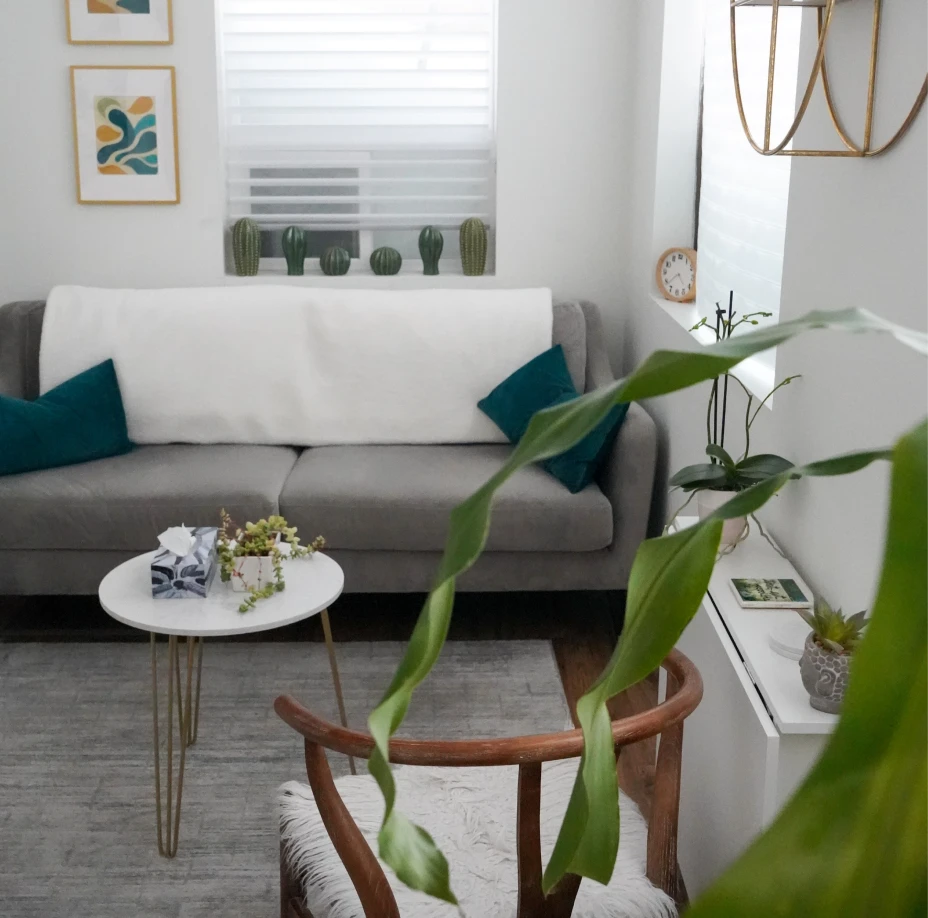 Modern therapy room with gray sofa, white throw blanket, green pillows, small round table with plant and tissues, wooden chair, and large green plant in foreground.