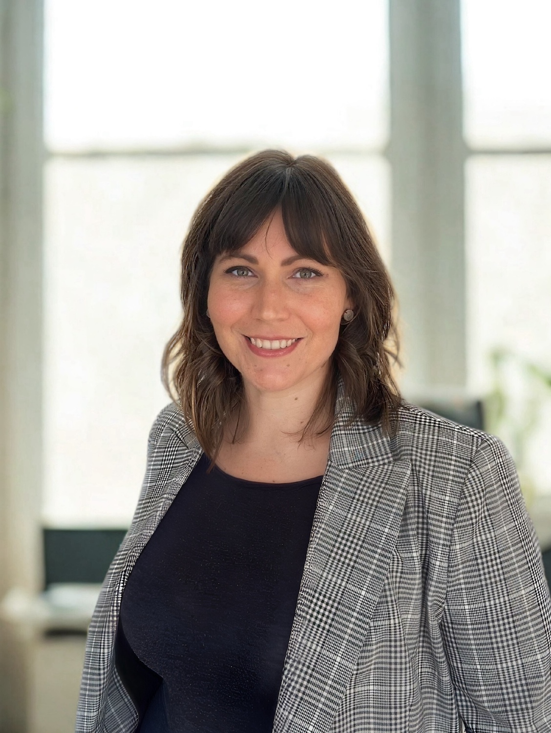 Sarah is standing with in front of a window smiling and facing the camera. She is wearing a houndstooth style blazer and black top. She has mid length brown hair. 