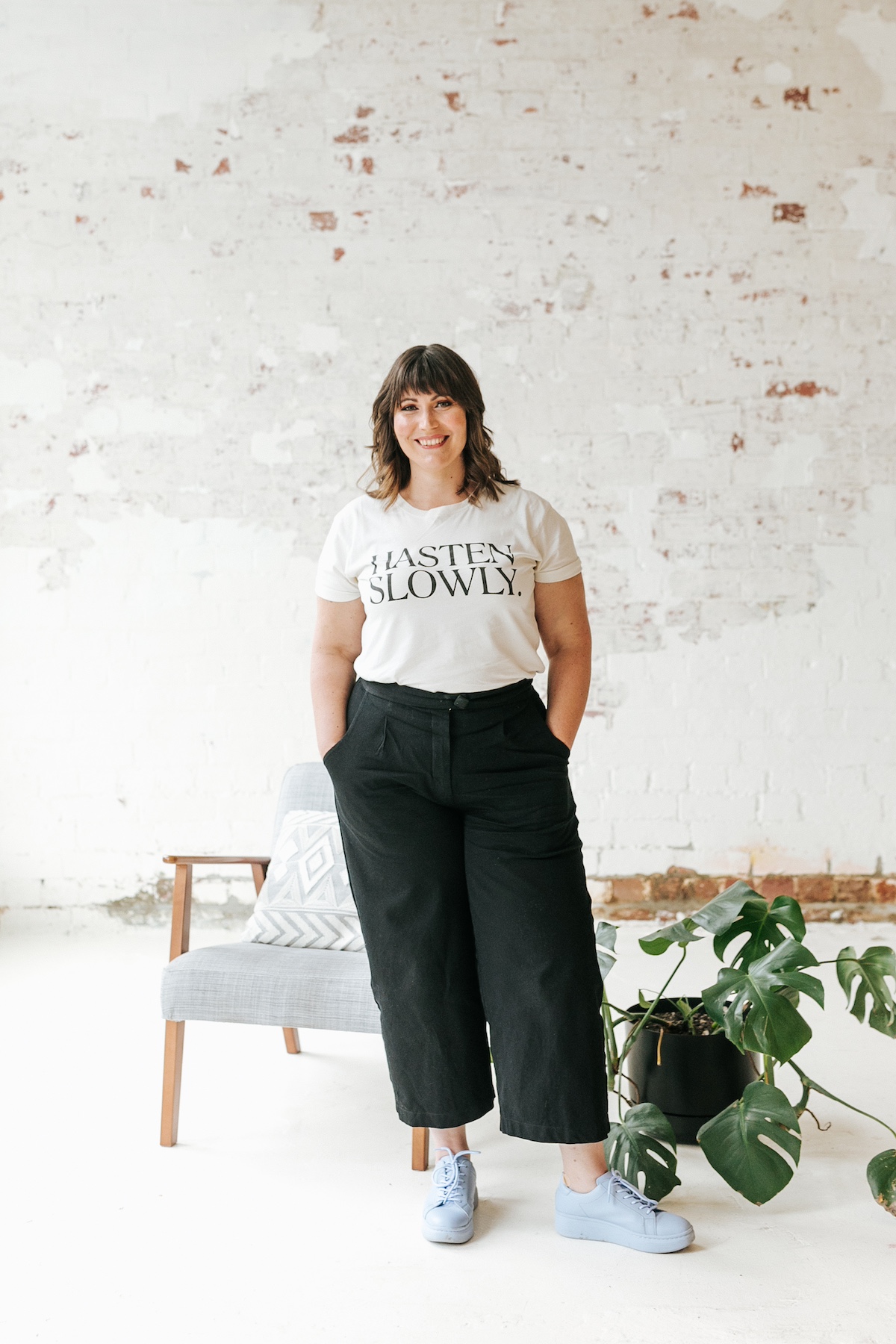 Sarah is standing in a bright room infront of a grey arm chair and rambling monsteria plant. She is wearing a white t-shirt that says "hasten slowly". She is smiling facing the camera.