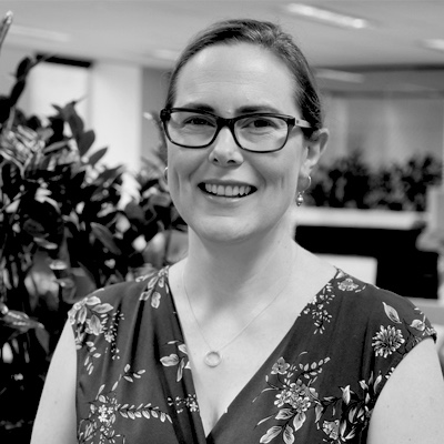 A black and white photo of Jacquie, smiling and facing the camera. She has brown hair tied back and glasses on. She's wearing a patterned top. An office plant is in the background.