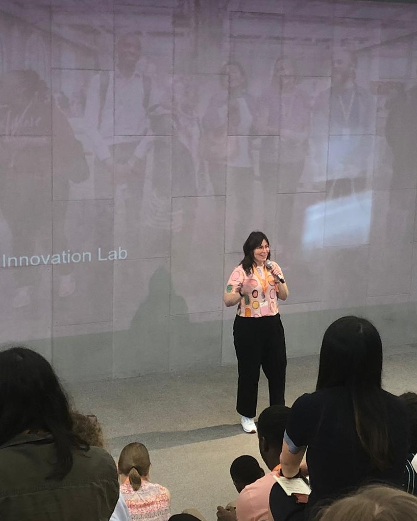 Sarah is standing in a bright room infront of a grey arm chair and rambling monsteria plant. She is wearing a white t-shirt that says "hasten slowly". She is smiling facing the camera.