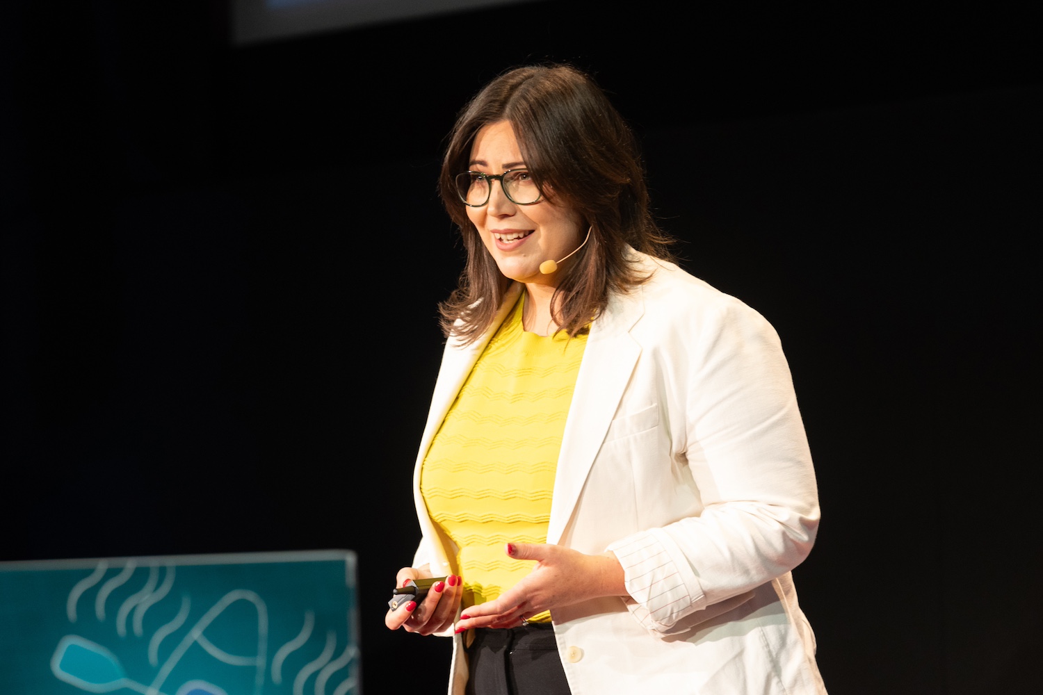 Sarah is addressing an audience with a serious look on her face. Her hands are open. She has a microphone and she is wearing a cream blazer and yellow top. You can see her body from the waist up. She is wearing glasses.