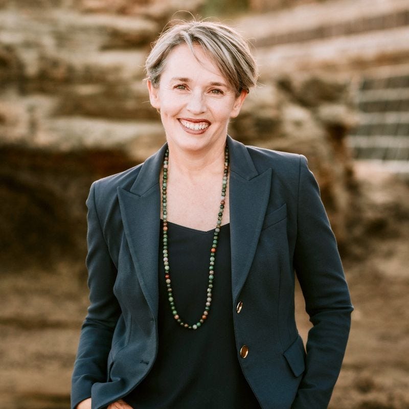 Michelle is standing smiling at the camera. She' has short blond hair and is wearing a dark green blazer. In the background there is a beautiful sandy rock.