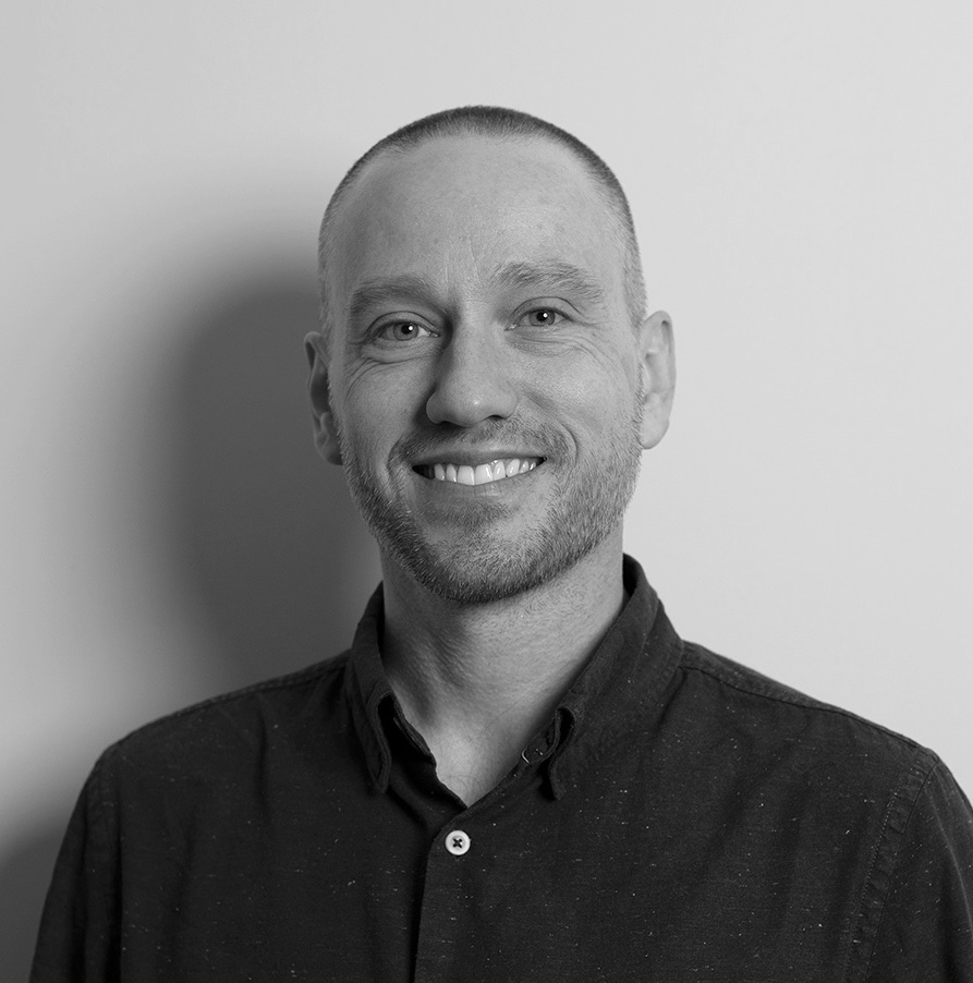 A black and white headshot of Cam. He is smiling and facing the camera. He has short, shaved hair and is wearing a dark collared shirt. His background is plain white.