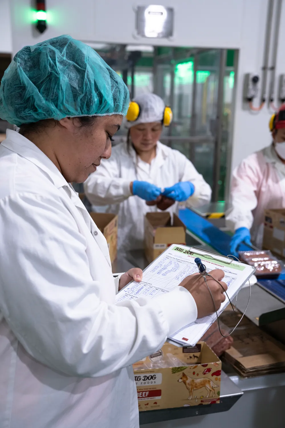 A Big Dog Pet Food factory worker in white coat and hairnet checking quality and recording data on a clipboard in the Big Dog Pet Food production line.