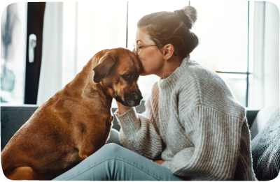 A woman on a couch cuddles a sad looking dog.