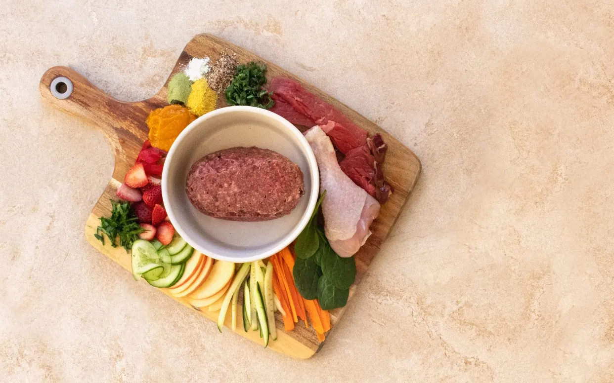 Raw patty in white bowl on chopping board surrounded by fresh ingredients including chicken meat, red meat, spinach, carrot, cucumber, apple and strawberries.