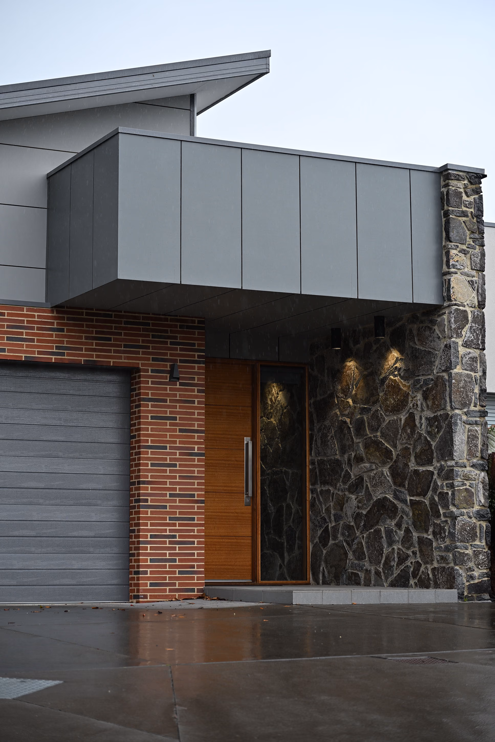 Modern house entrance with gray garage door, wood door, brick and stone walls, and exterior lighting.