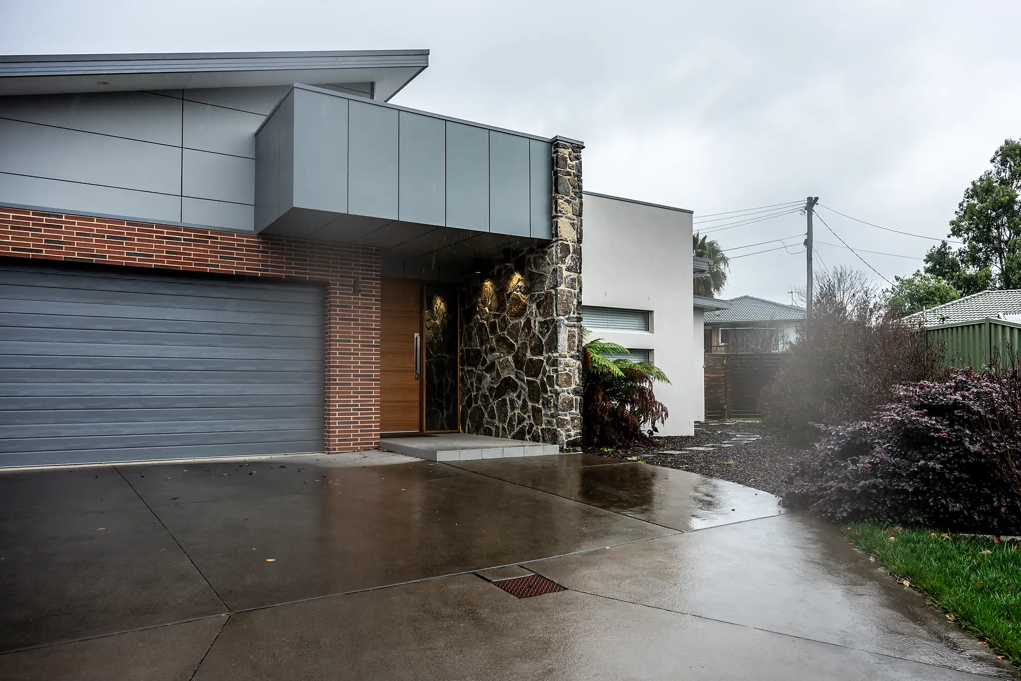 Modern house exterior with stone, brick, and gray garage door on a wet driveway under overcast sky.