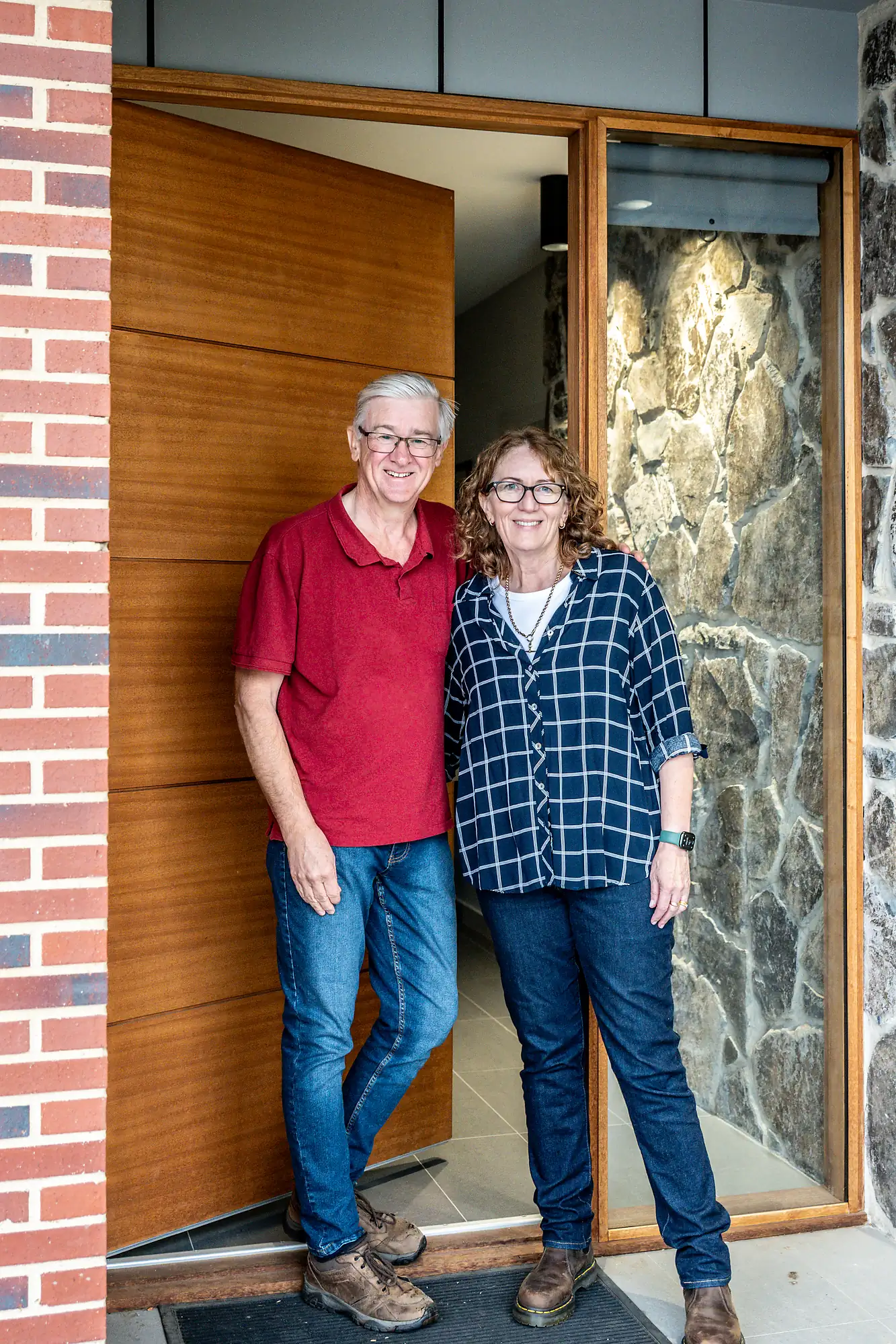 Smiling couple standing at the entrance of a house with a wooden door and stone walls.