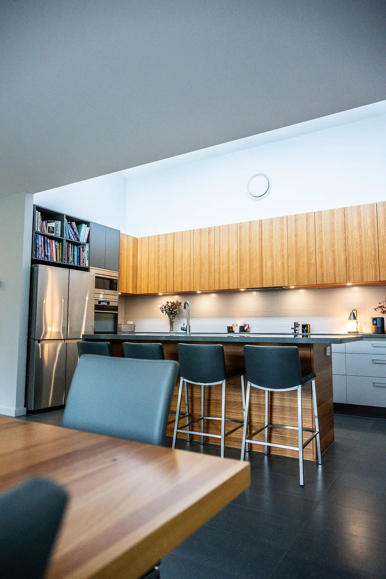 Modern kitchen with wood cabinets, stainless appliances, gray bar stools, and a wooden dining table.