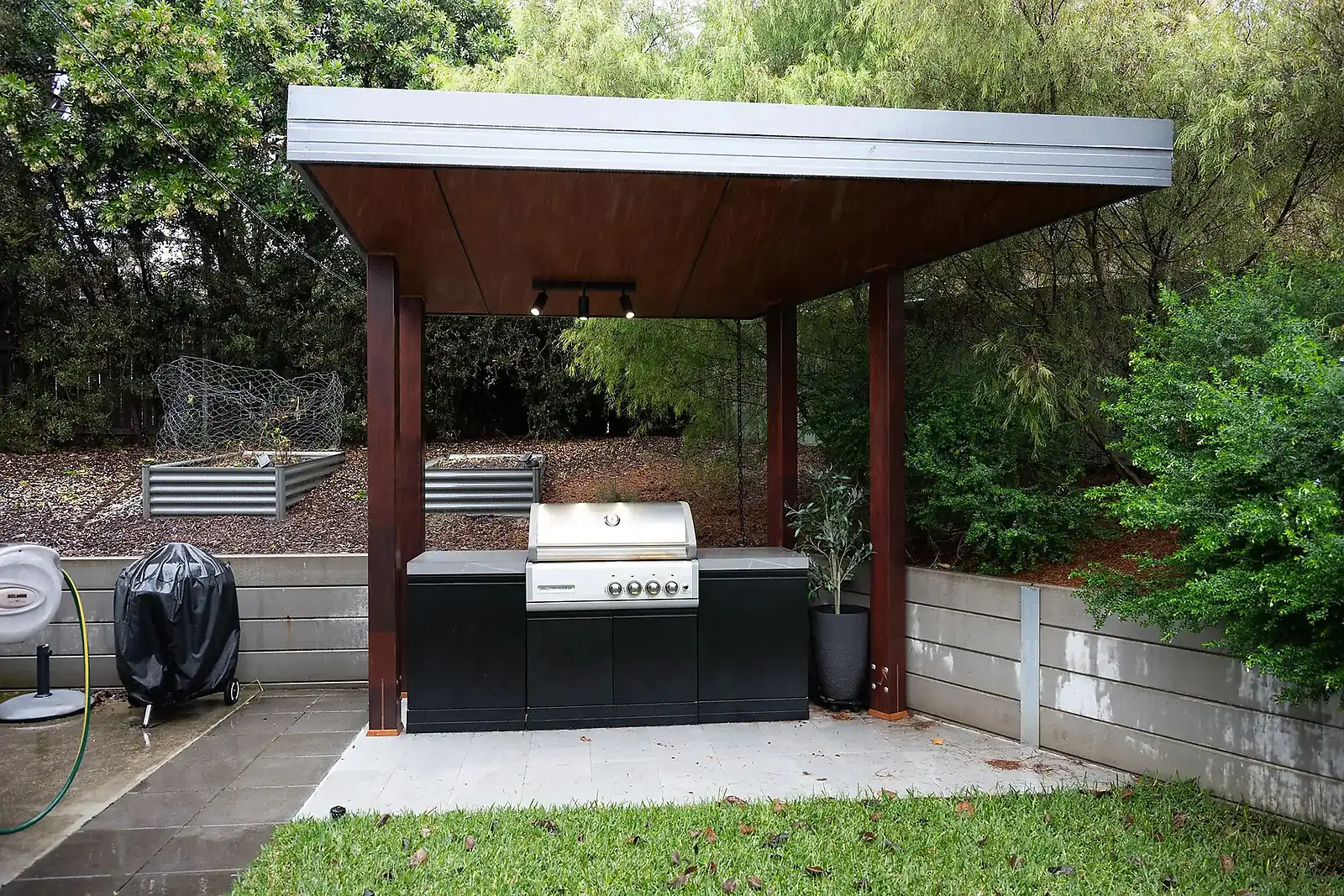 Outdoor covered grill station with stainless steel grill, black cabinets, plants, and garden beds in the background.