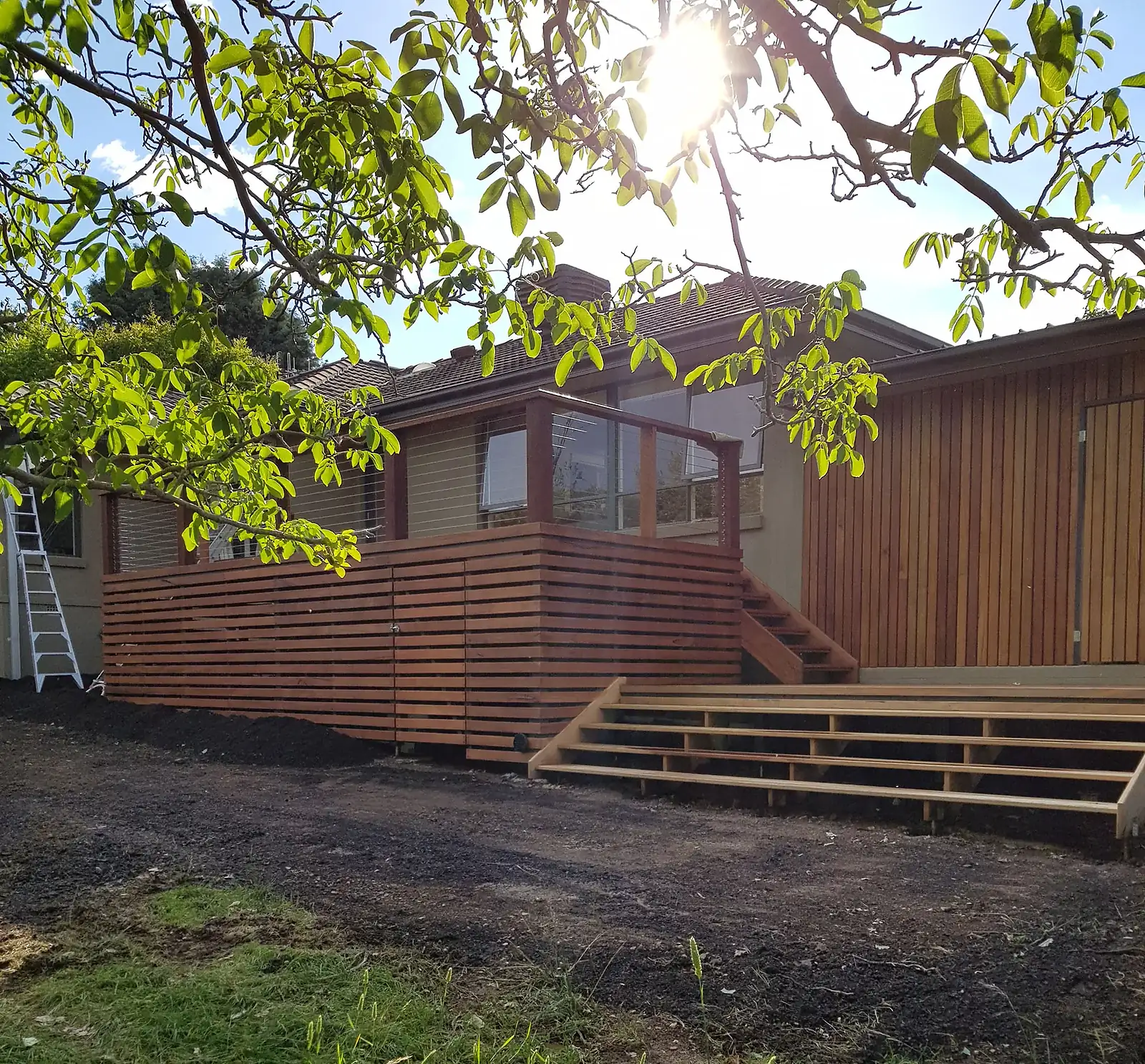 Modern house with a wooden deck, steps, and railing under tree branches in sunlight.