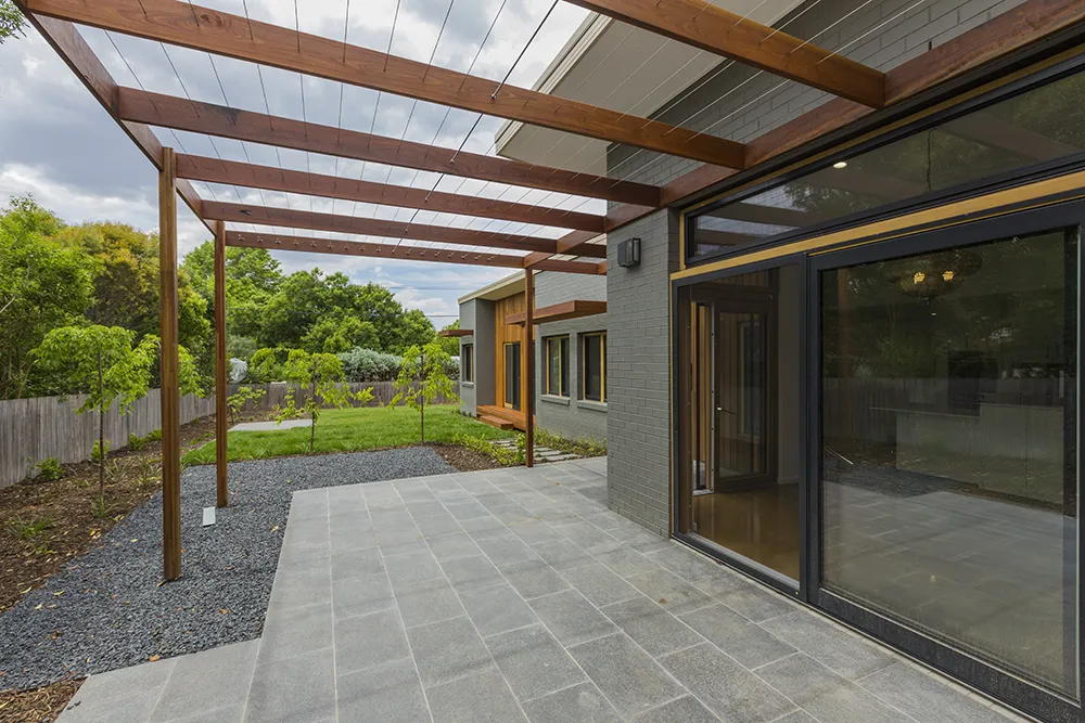 Modern patio with gray tiles, wooden pergola, sliding glass door, and green backyard with young trees.