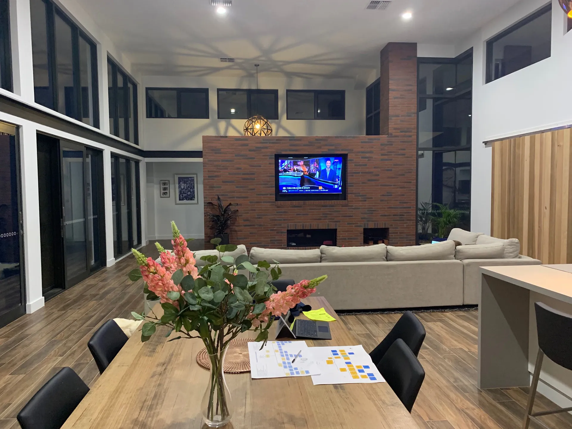 Modern living room with large beige sofa, brick TV wall, wooden floor, and flowers on a dining table.