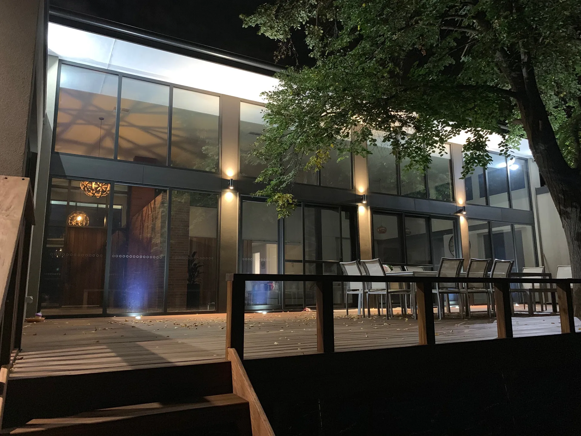Outdoor patio at night with wooden deck, chairs around tables, and a tree on the right side.