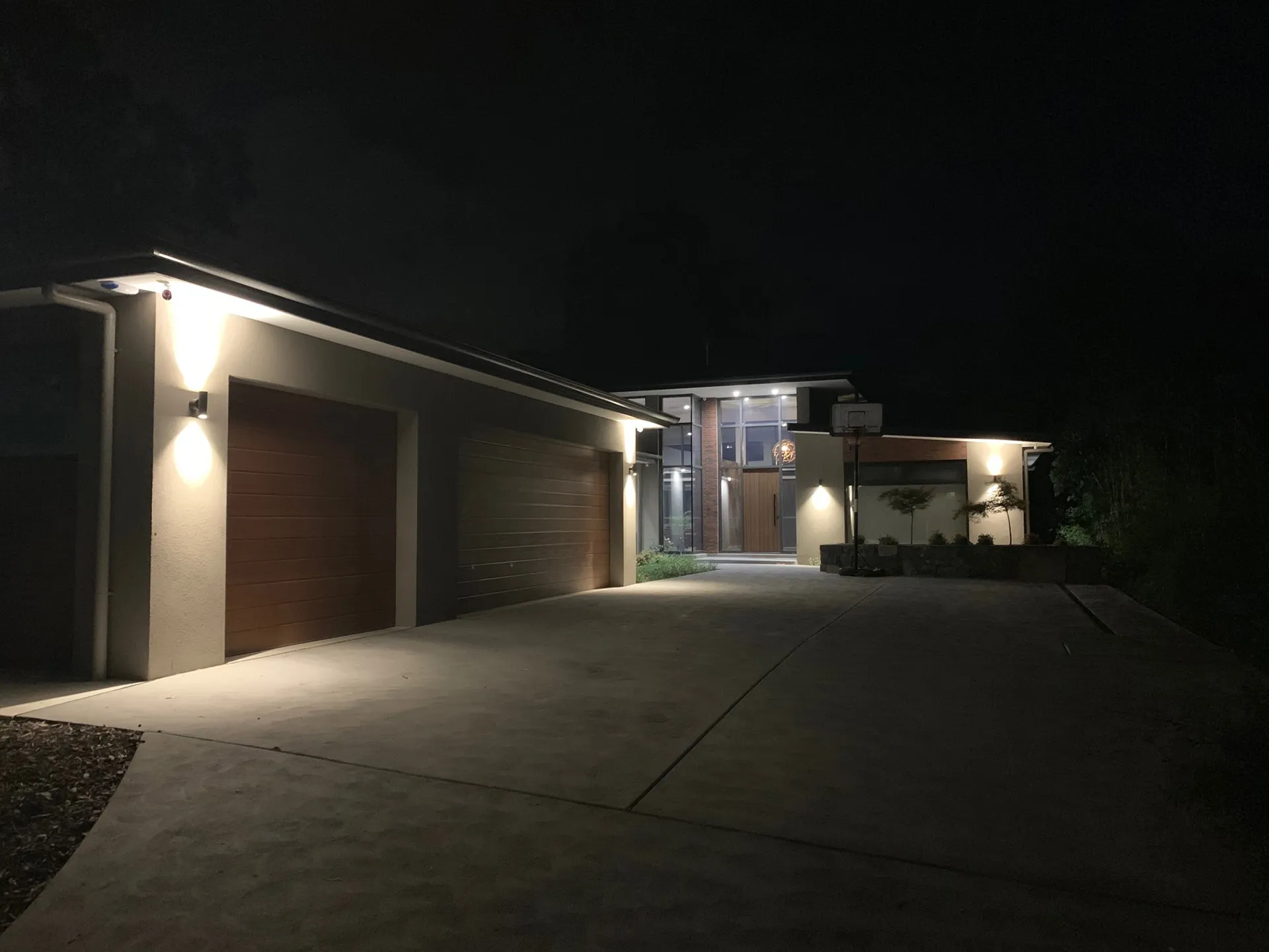 Modern house driveway at night with exterior lights highlighting garage doors and entrance.