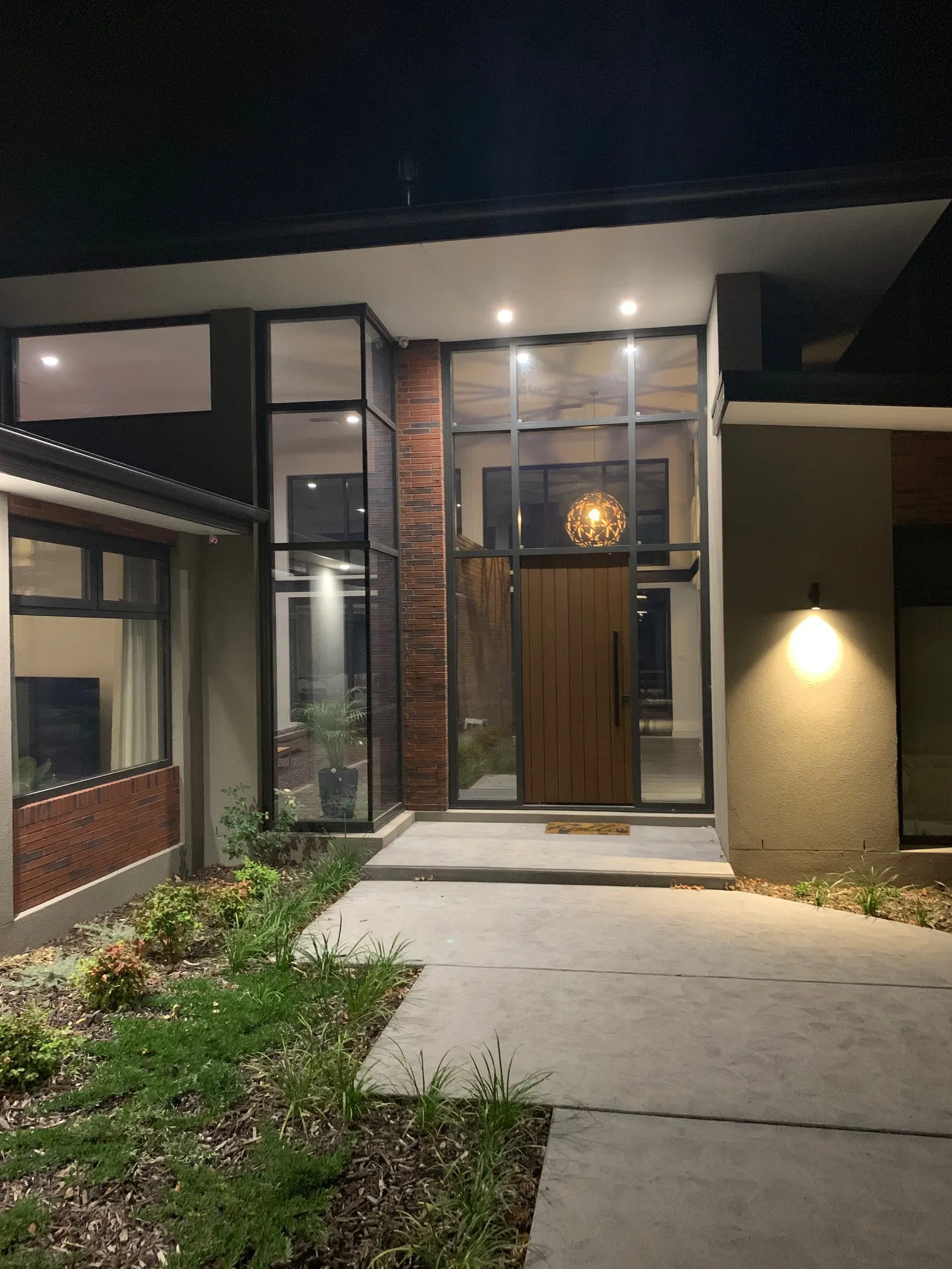 Modern house entrance at night with glass walls, a wooden door, and outdoor lighting.
