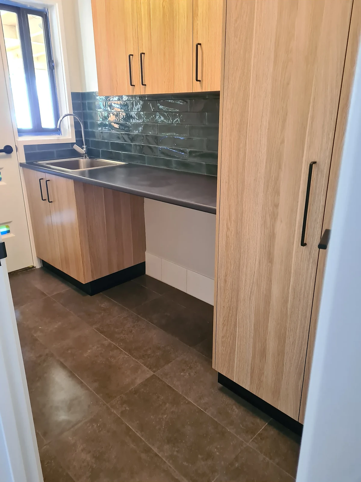 Compact kitchen with wooden cabinets, dark countertop, green tile backsplash, and stainless steel sink.