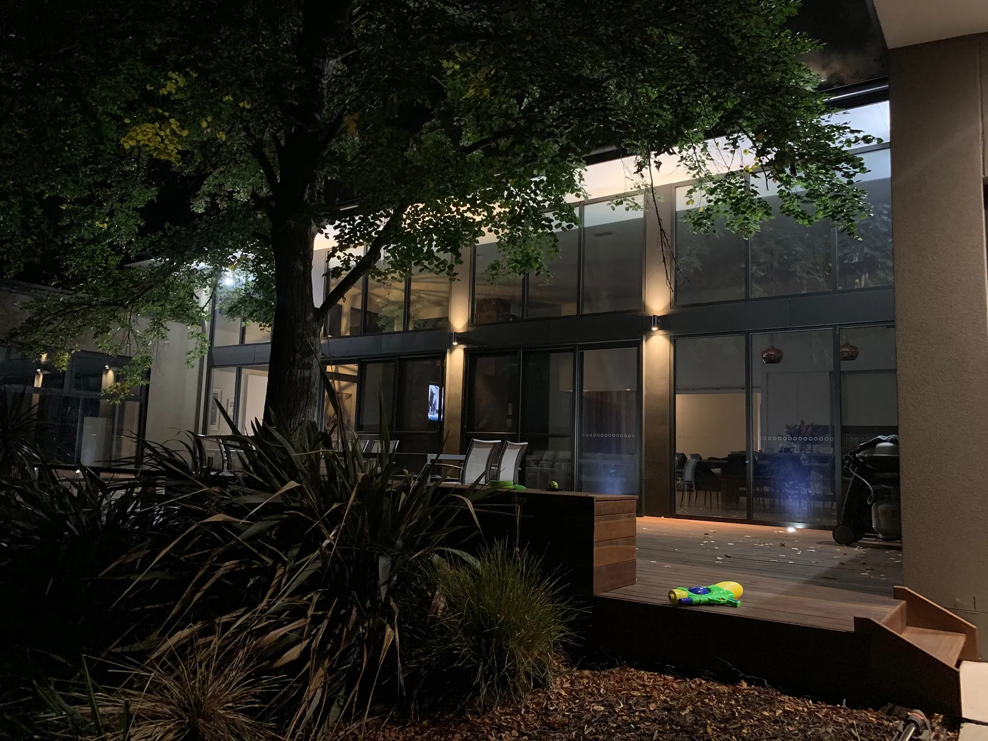 Night view of a modern house patio with chairs, plants, a tree, and a toy water gun on the wooden deck.