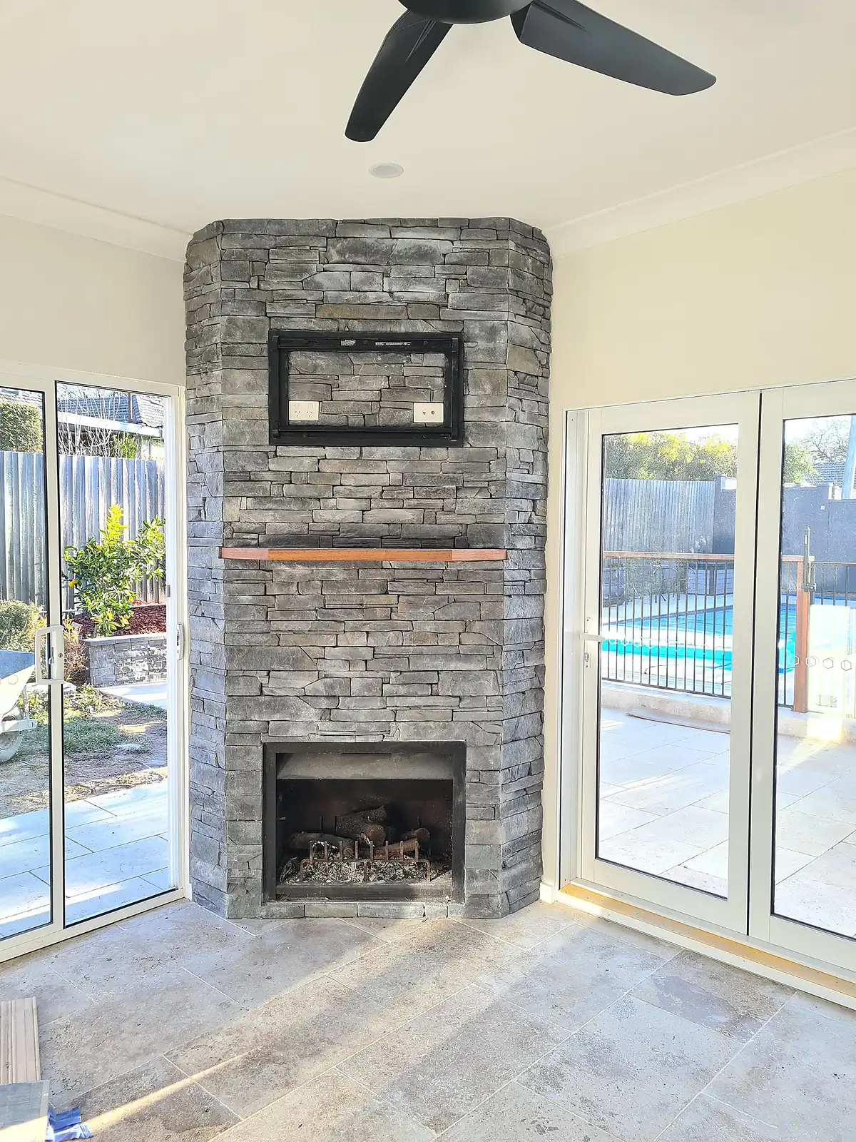 Stone fireplace with a wooden mantel in a room with sliding glass doors showing a pool outside.