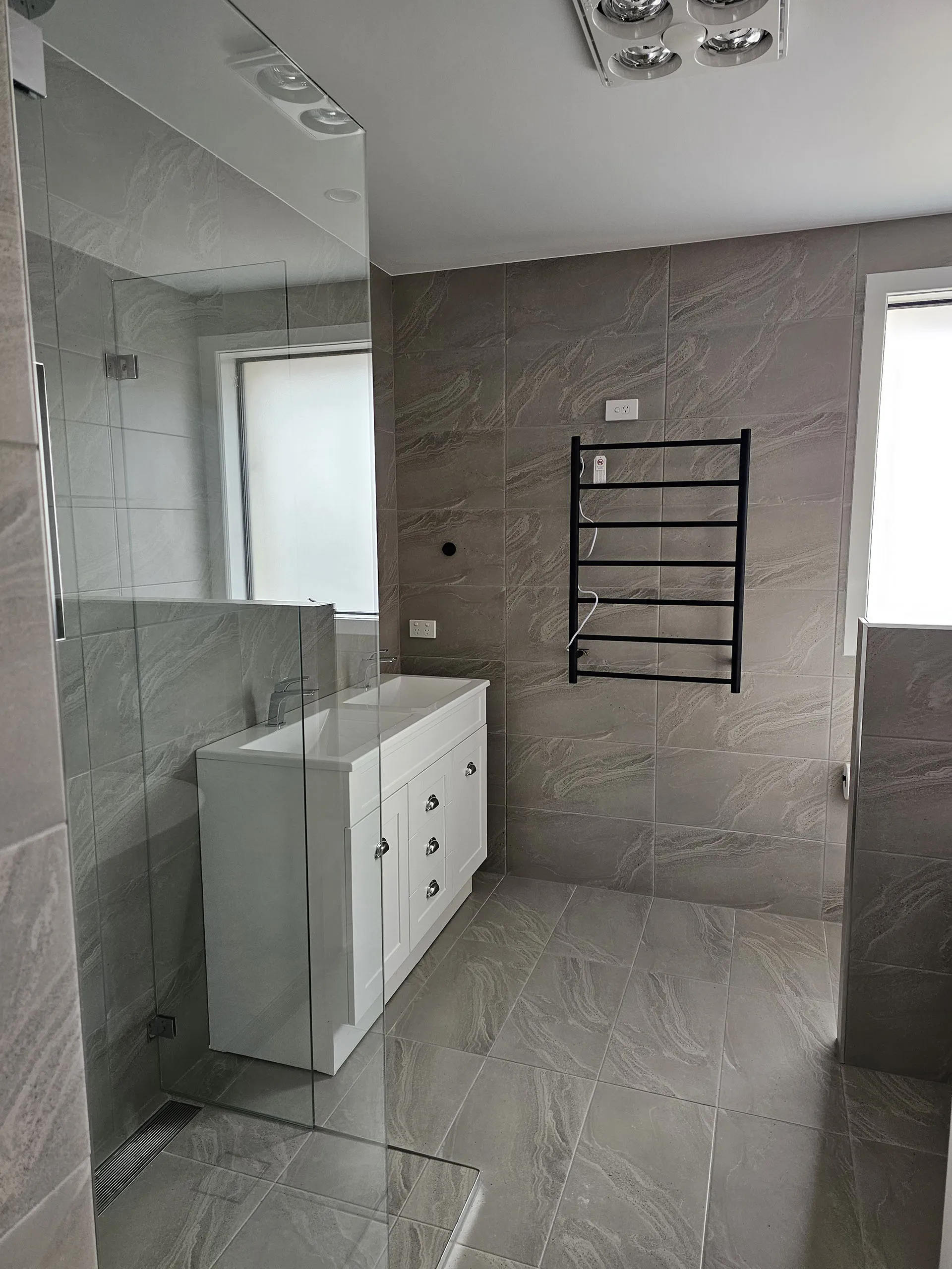 Modern bathroom with gray marble tiles, white vanity, glass shower, and black towel rack on wall.