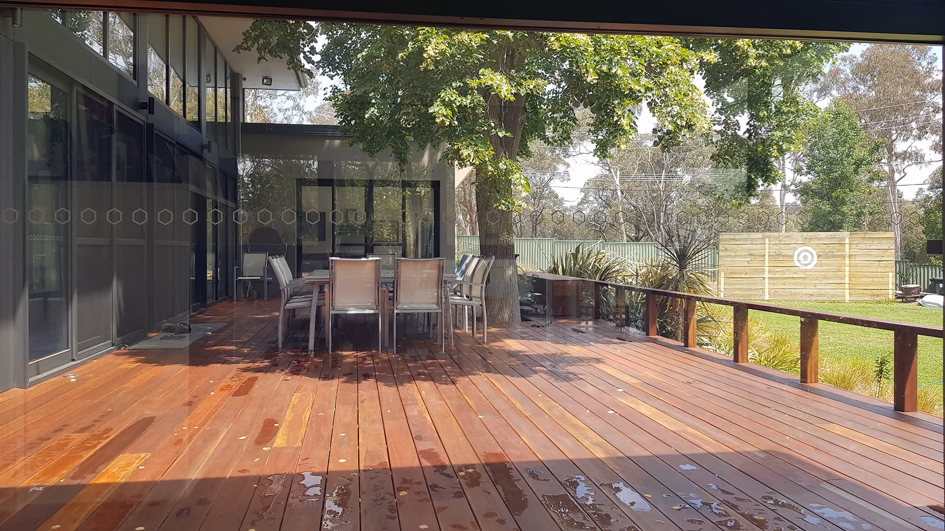 Outdoor wooden deck with dining table, chairs, large tree, and green yard with target on wooden fence.