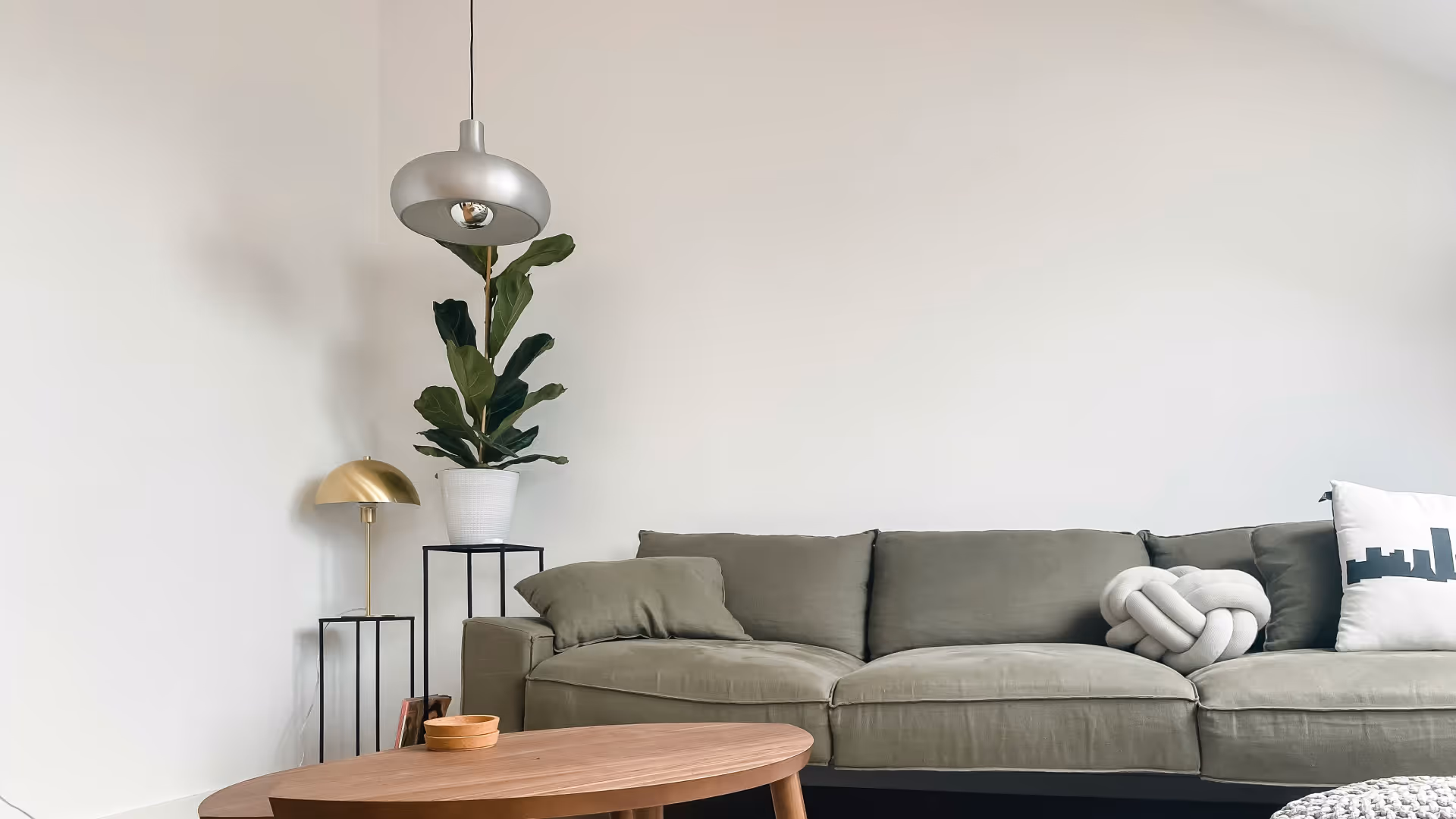 Minimalist living room with a green sofa, wooden coffee table, tall plant in a white pot, and modern pendant and table lamps.