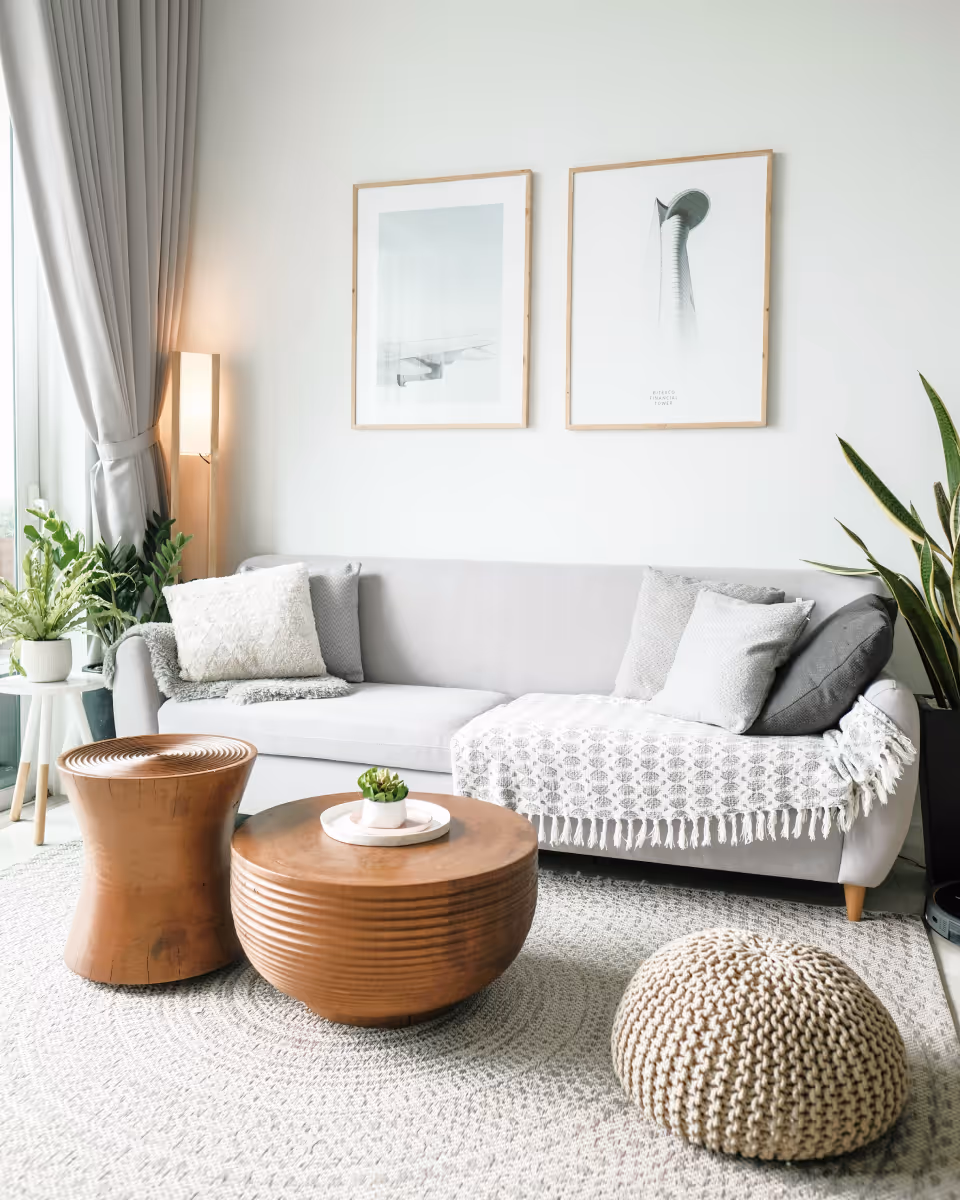 Modern living room with a light gray sofa, wooden coffee tables, knitted pouf, plants, and minimal wall art.