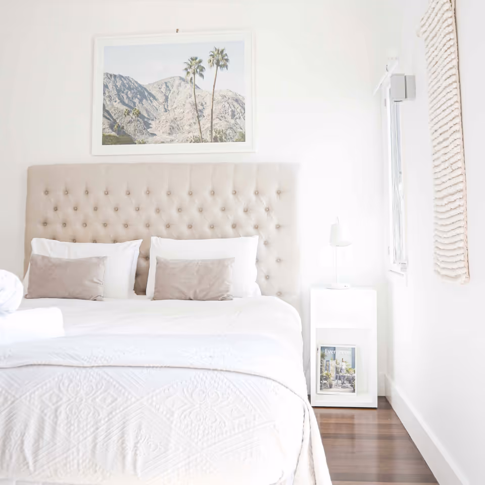 Bright bedroom with a tufted beige headboard, white and beige pillows, white bedding, a nightstand with a candle and magazine, and a framed mountain and palm tree photo on the wall.