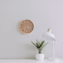 Minimalist workspace with a small potted plant on a desk, a white desk lamp, and a simple round clock on a white wall.