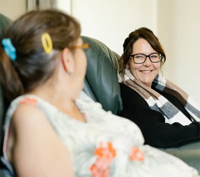Two women sitting on a green couch, one smiling and wearing glasses and a striped scarf, the other with a hair clip and glasses, blurred in foreground.