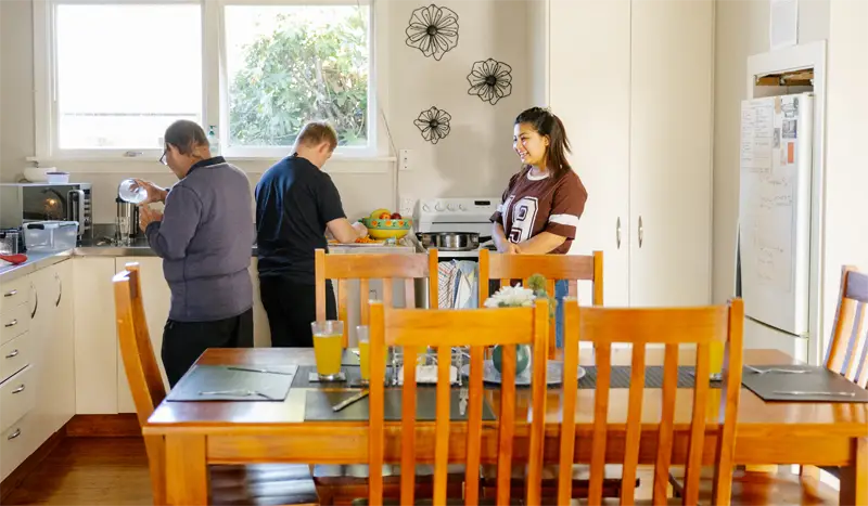 Three people engaged in kitchen activities behind a wooden dining table set with placemats and a flower vase.