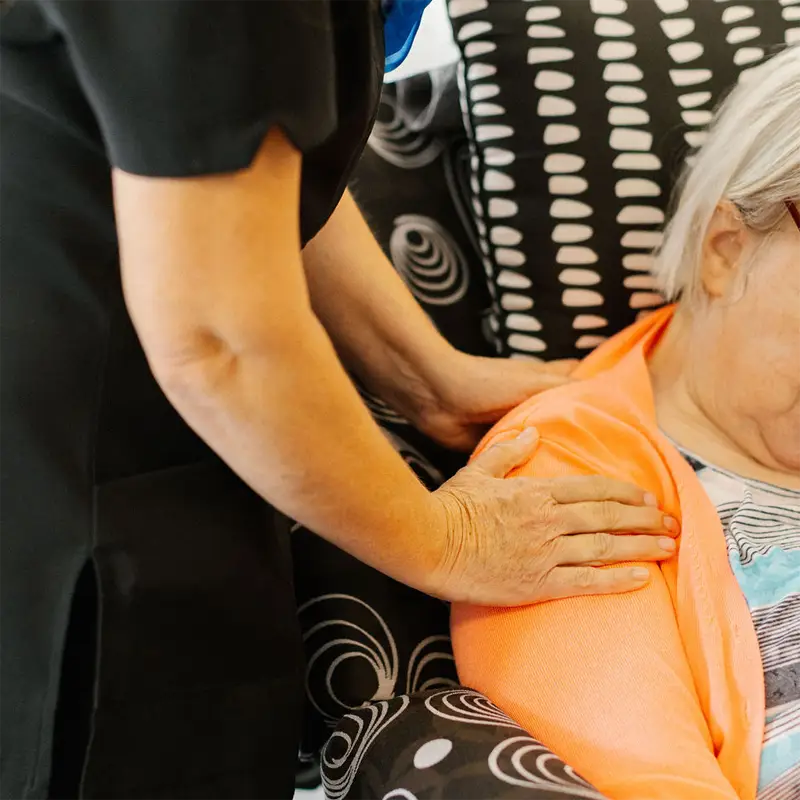 A caregiver wearing black gently massages the shoulder of an elderly woman seated on a patterned chair.