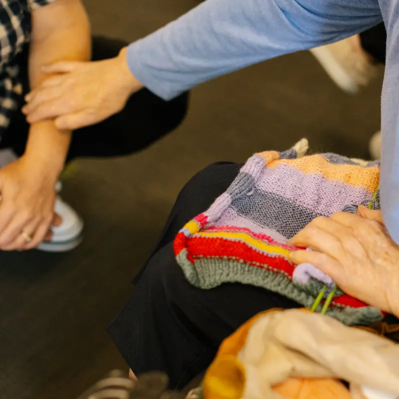 Hands of two people, one gently holding the other's arm, with a colorful knitted blanket resting on a lap.