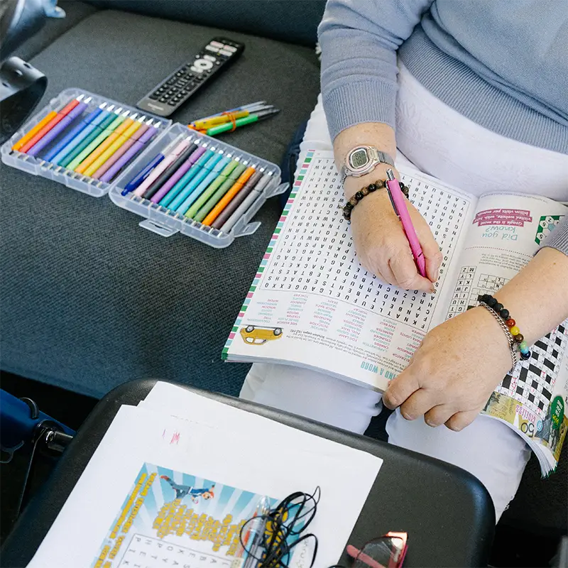 Person wearing a watch and bracelets doing a word search puzzle with a pink pen, colored pencils and pens on a gray couch nearby.