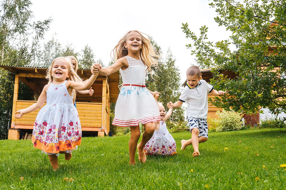 Four children holding hands and running barefoot on green grass in a backyard with wooden buildings and trees.