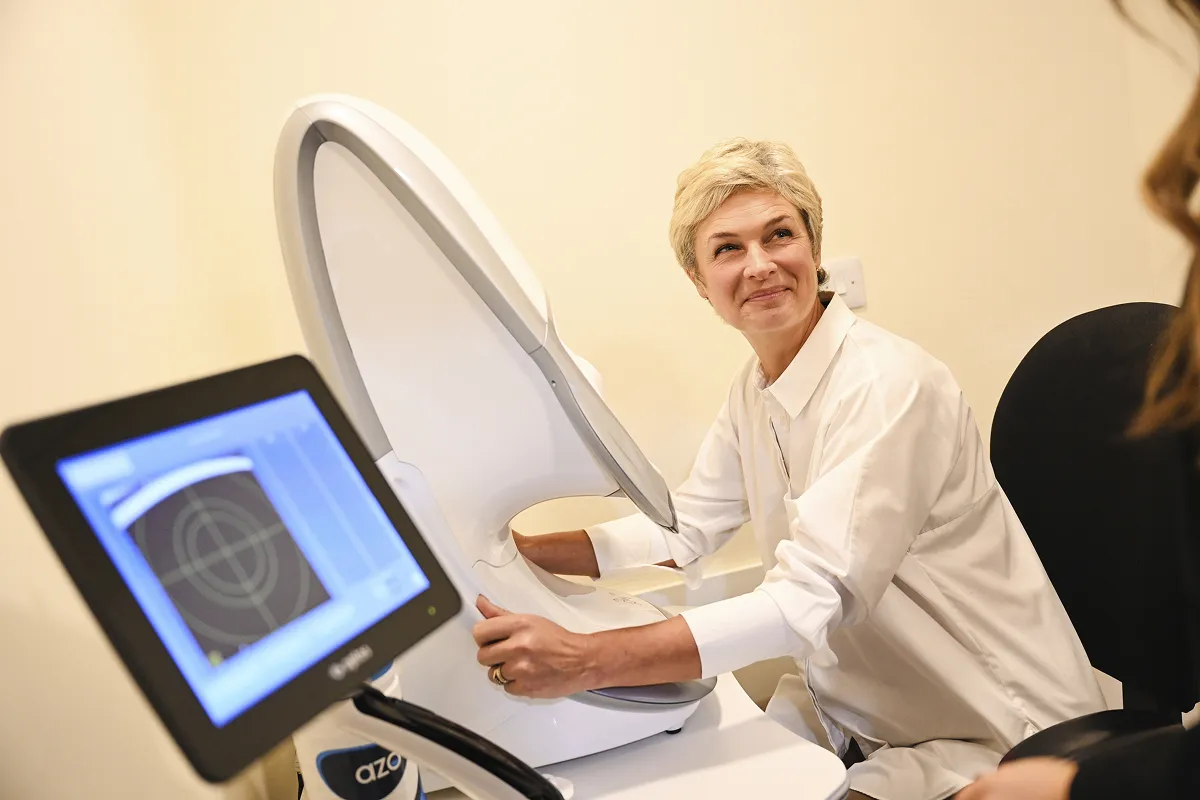 A woman in a white lab coat operates a machine with a screen displaying a target target pattern, while smiling and looking toward a person outside the frame.