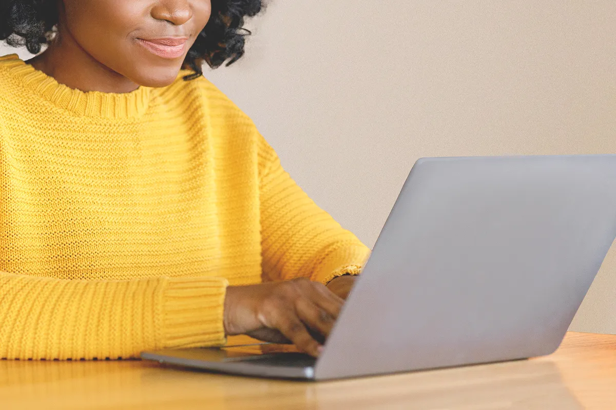 Person in a yellow sweater typing on a laptop at a wooden table.
