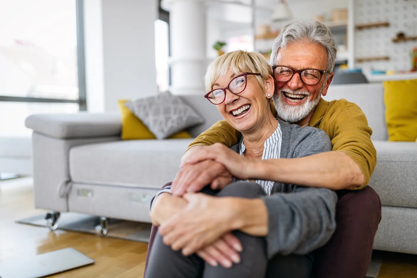 Smiling elderly couple wearing glasses embracing while sitting on the floor in a bright living room with a gray couch and yellow pillows.