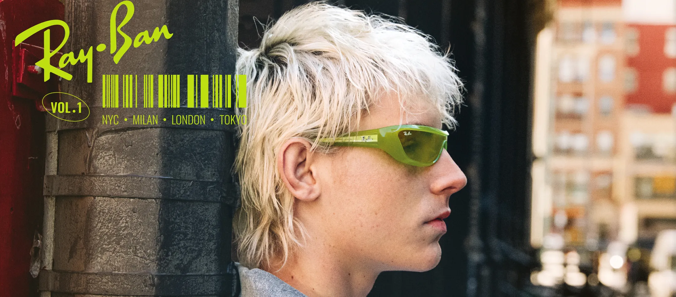Profile of a person with platinum blonde hair wearing green Ray-Ban sunglasses leaning against a textured wall in an urban setting.