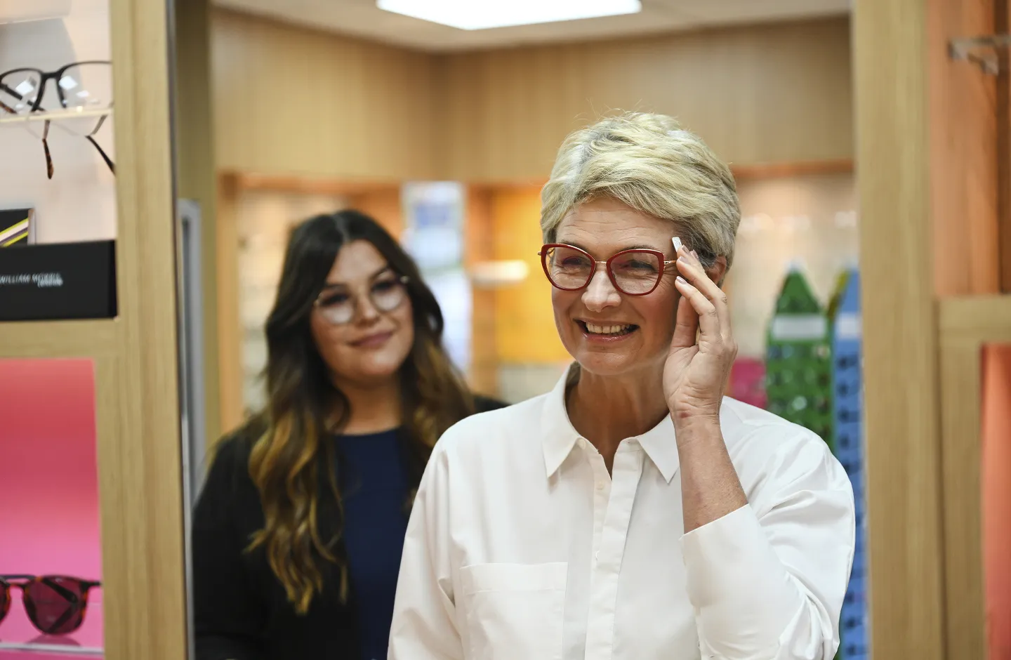 Smiling older woman trying on red eyeglasses with a younger woman watching in an eyeglass store.