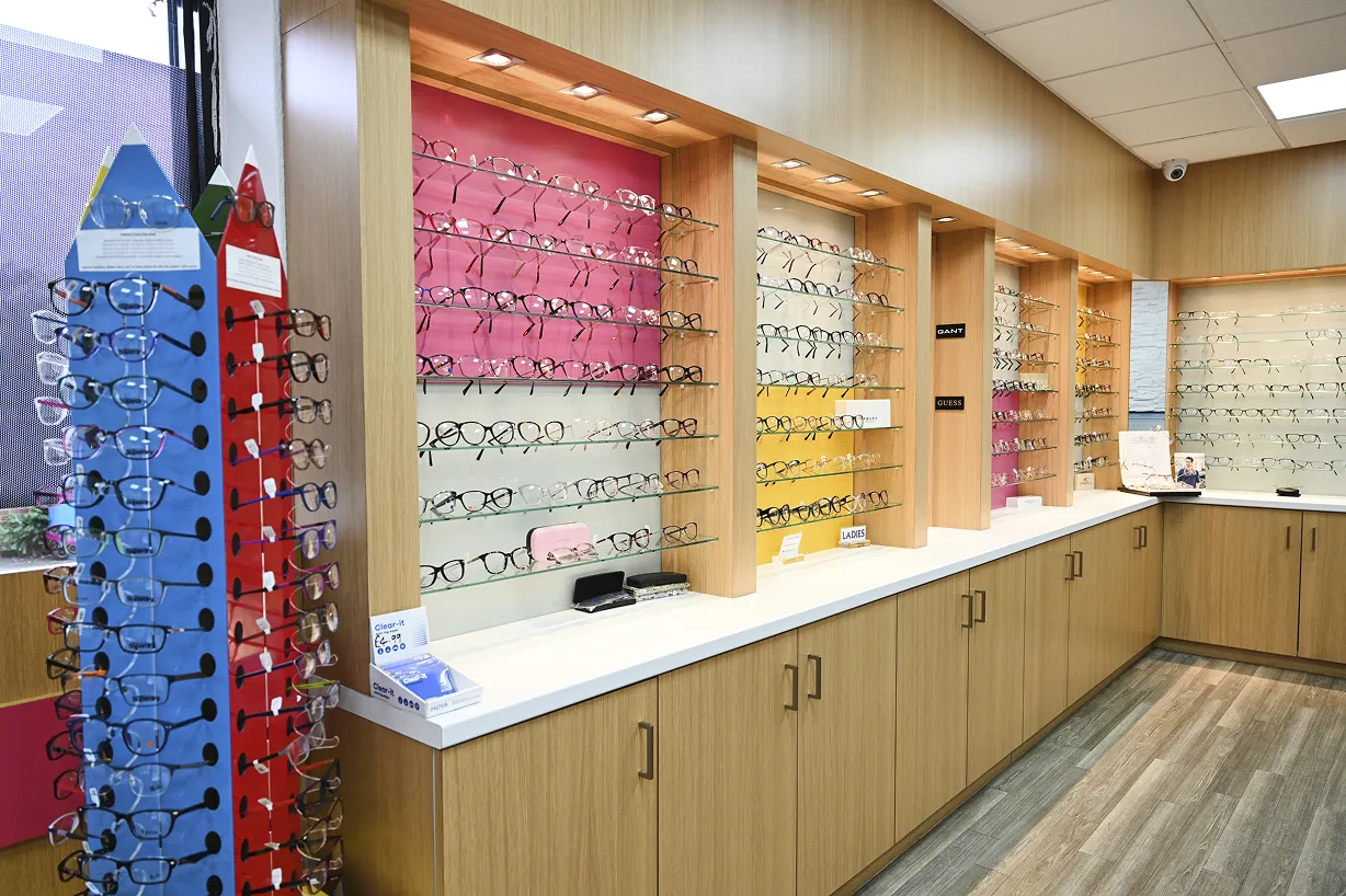 Interior of an eyewear store with multiple shelves displaying various eyeglass frames and a free-standing blue and red rack of glasses near a window.