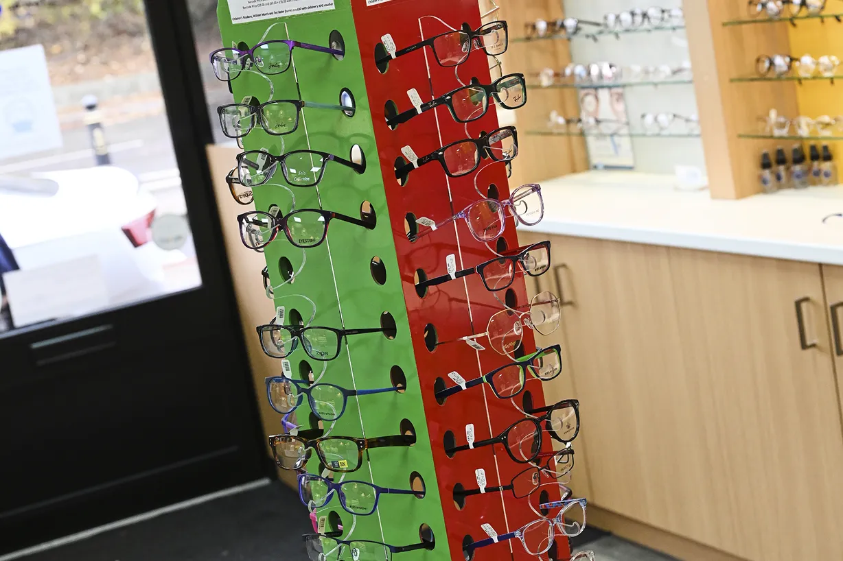 Display rack with various eyeglasses frames inside an optical store near a counter and shelves.