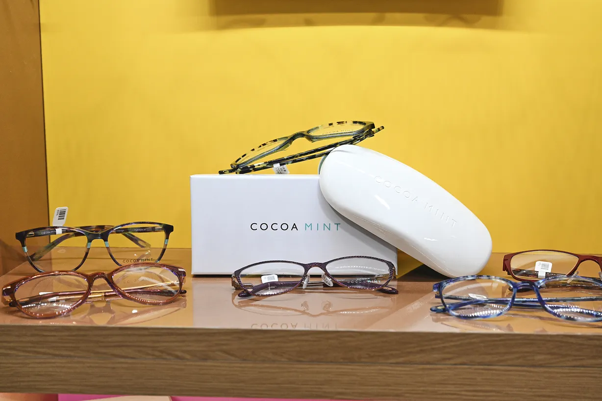 Display of five pairs of eyeglasses with varied frame colors arranged on a wooden surface next to a white box and glasses case labeled 'Cocoa Mint'.