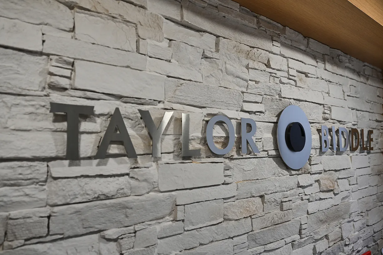 Metal letters spelling 'TAYLOR BIDDLE' mounted on a textured light gray stone wall with a round logo between the words.