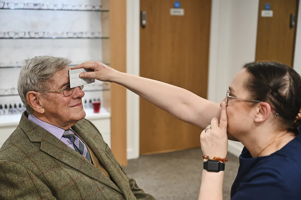 Optometrist holding eyesight test card to an elderly man's forehead during an eye exam in an optical store.