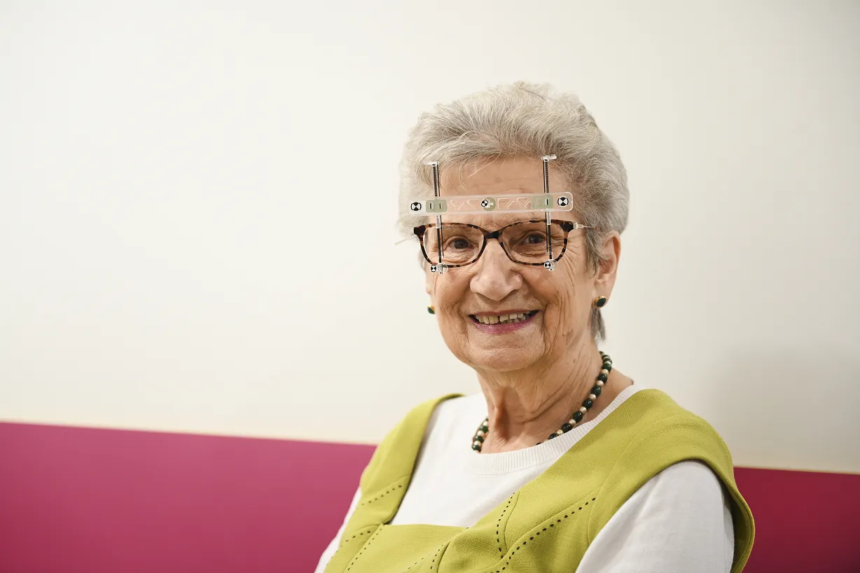 Smiling elderly woman with gray hair wearing glasses and a green vest over a white shirt, sitting against a white and pink wall.